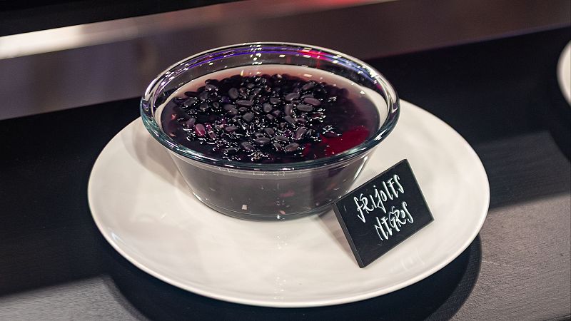 Frijoles negros cocidos en un cuenco de cristal sobre un plato blanco, identificados con un cartel que dice "Frijoles Negros".